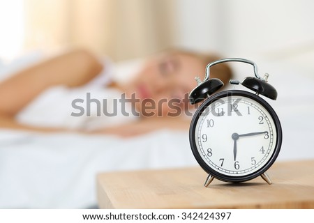 Alarm clock standing on bedside table going to ring early morning to wake up woman in bed sleeping in background. Early awakening, not getting enough sleep, oversleep, getting work time concept
