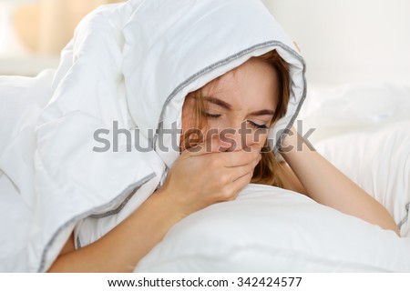 Sick young woman lying in bed suffering with cold covering nose with hand while sneezing. Female feeling sickness and unwell hiding head under blanket. Pregnancy, period premenstrual syndrome concept