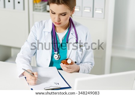 Beautiful female physician medicine doctor or pharmacist sitting at worktable, holding jar of pills in hands and writing prescription on special form. Medical care, pharmacy or insurance concept