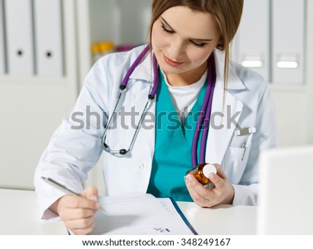 Beautiful female physician medicine doctor or pharmacist sitting at worktable, holding jar of pills in hands and writing prescription on special form. Medical care, pharmacy or insurance concept