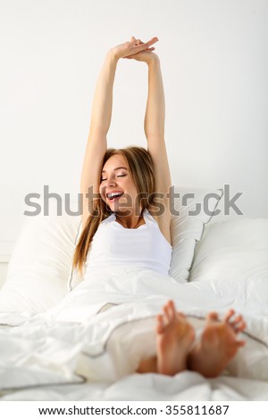 Young beautiful smiling and yawning blonde woman sitting in bed trying to wake up early morning stretching hands and legs after sleep. Sweet dreams, good morning, new day, weekend, holidays concept