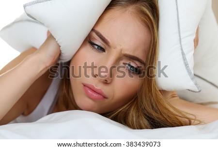 Young beautiful woman lying in bed suffering with insomnia covering head and ears with pillow and making unpleasant face. Noisy neighbour, stress, alarm sound, prevent from sleep concept