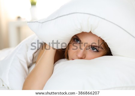 Young beautiful woman lying in bed suffering with insomnia covering head and ears with pillow and making unpleasant face. Noisy neighbour, stress, alarm sound, prevent from sleep concept