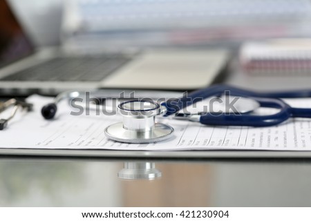 Stethoscope head lying on medical form on clipboard pad at empty doctor worktable with laptop computer. Physical, disease prevention, examine patient, 911, instrument shop, healthy lifestyle concept