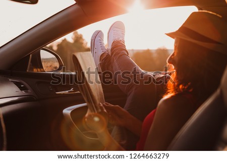 Modern hipster girl resting in a car and reading a map. Woman with feet on car door. Feet outside the window at sunset