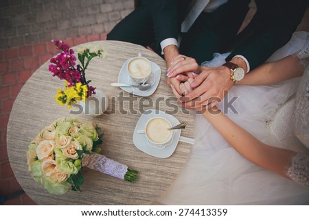 Cosy cafe wedding bouquet lying on the table. Bride and groom sitting at the table, having coffee and holding hands