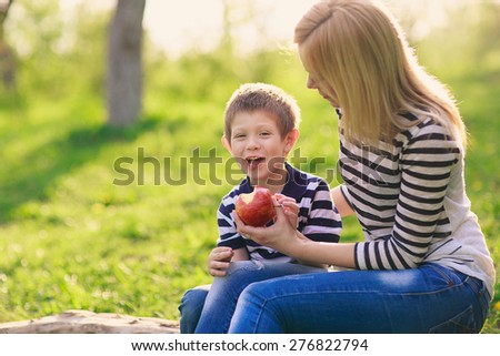 happy mother and her young son sitting on a tree in the park and eat a big red apple