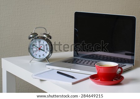 alarm clock, note book with black pen on it, red cup of coffee and silver laptop on the table