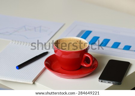 a red cup of coffee, business diagrams, mobile phone, note book and black pen on the table