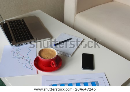 a red cup of coffee, laptop, business diagrams, mobile phone, note book and black pen on the table