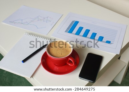 business graphs and diagrams, note book, pen, cellphone and red cup of coffee on the table