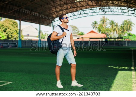 Young stylish men posing on a soccer field,listen music.Having fun.Wear cool sportswear,big sport bag,denim shorts,summer reflecting sunglasses,a young man with beard,football fan,Listen music