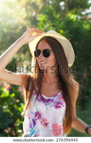 Close-up portrait of young woman in park wear stylish straw hat,sunglasses and fashionable summer clothes,tanned and beautiful model on vacation,tropical summer mood,flower print,summer portrait