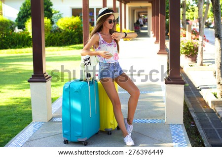 Young woman with luggage at airport,look at her watch,waiting departure.tanned girl with large suitcases,straw hat,sweater,summer sunglasses,denim shorts,happy stylish hipster girl,vintage teen style