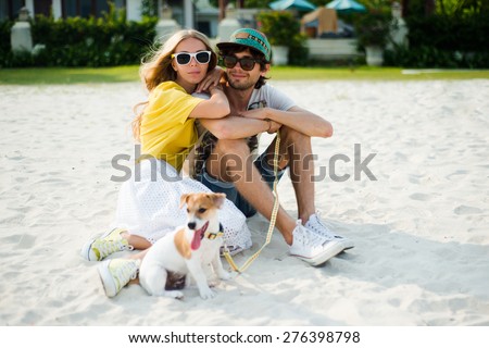 Young happy couple in love sitting on the beach in summer sunny evening, with dog. Summer mood,smiling and having fun during their vacation, wearing sunglasses, cap, yellow and printed shirt, romance