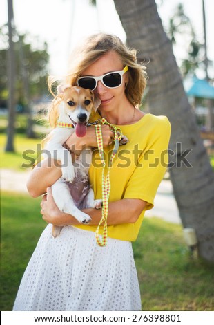 Sunny summer portrait of stylish hipster girl holding a dog at the park,smiling and having fun,wear summer sunglasses,vintage clothes,yellow shirt and white skirt,tropical background,sunny colors