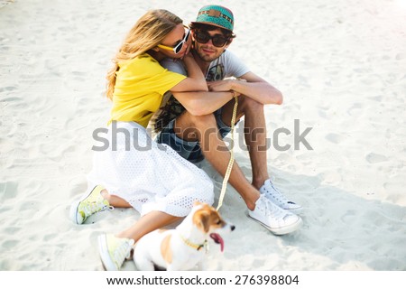 Fashionable couple in love sitting on the beach of white sand, playing with puppy. Summer close-up couple in love,wear summer sunglasses,cap,yellow shirt,fashion lifestyle store,glowing skin