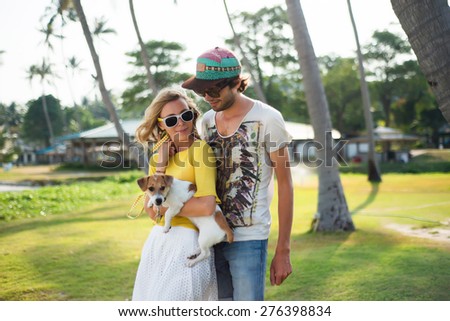 Hipster couple in love on the beach in summer sunny evening,with dog.Summer mood,summer beach portrait of young couple in love,wearing sunglasses, cap, yellow shirt, island romance.Palms on background