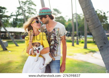 Young couple in love on park opposite the palm trees, summer sunny evening,with dog. Summer mood,summer beach portrait of young couple in love,wearing sunglasses, cap, yellow shirt, island romance