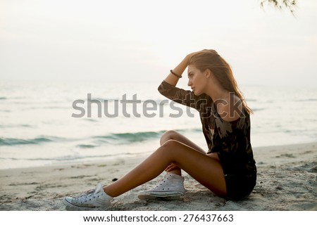 Young girl sitting on the beach after sunset in the sea background.Young traveler.Wear clothes for a hike,camouflage Sweatshirt and sneakers.Dreaming,summer denim clothes,tan girl,