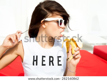 Young pretty teen girl with wearing stylish hipster outfit, and eating banana.\
Party concept.posing on the beach resort background, bright summer fashion portrait.
