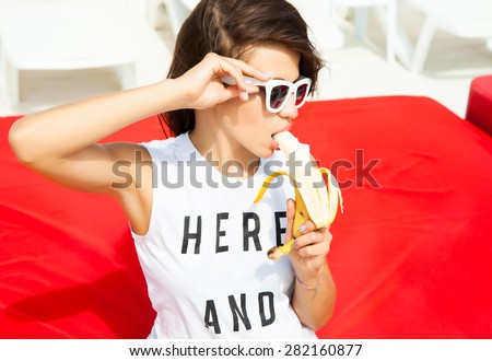 Young pretty teen girl with wearing stylish hipster outfit, and eating banana.
Party concept.posing on the beach resort background, bright summer fashion portrait.