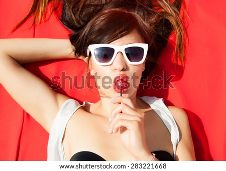 Happiness bliss freedom concept of sensual woman laying on red with summer sunglasses,eating sweets on bikini with glowing skin,having sunbath at hot sunny day.Summer outfit,fashion store,summer store