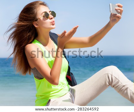 young woman with mobile phone on a beach.Sending kiss to her phone using front camera.Take Selfie pictures and taking white smartphone after workout. Healthy lifestyle female model in 20s.