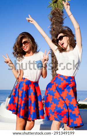 Two women friends laughing with a perfect white teeth with a green background,sea background,fashion store,beach party time,summer fashion store,trendy skirts,palm trees background,friend on vacation