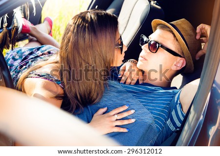 Romantic young couple sitting on the hood of their car while out on a road trip.Beautiful young woman kissing her boyfriend looking away smiling,outdoors.Holding hands and kissing at car,enjoy trip