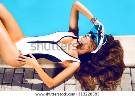 Beautiful sexy young girl with perfect slim figure with long wet hair and bathing suit in fashionable stylish sun glasses sitting on the steps of swimming pool swim, sunbathe, have fun at beach party