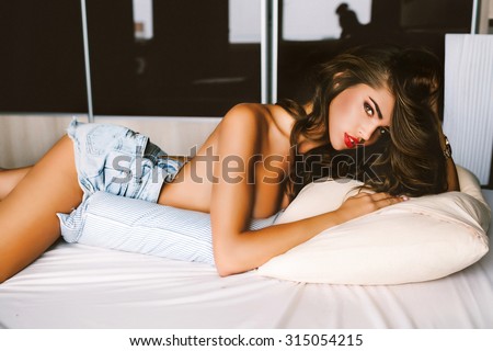 Soft photo of amazing woman with red lips,volume hair,curly hairstyle,denim shirts,glowing skin,big full lips posing at bedroom,wearing sexy luxury stylish.Lady lying on stomach on white bed,sexy tan