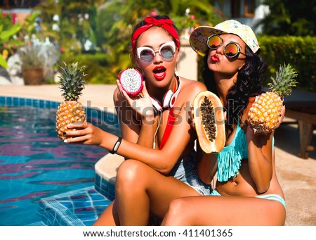Summer lifestyle happy smiling portrait of two pretty young women blonde and brunette fashion stylish girls having fun on the beach on tropic island in sunglasses and tropical fruits on their hands