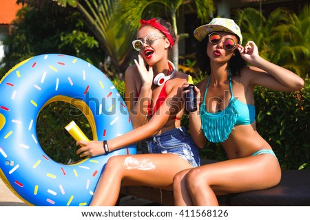 Summer portrait of beautiful stunning blonde and brunette happy girls sitting near the pool on the beach,and green palms background smiling and having fun together with sun cream,funny sunglasses,cool