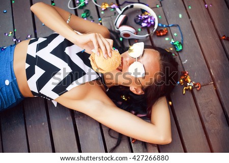 Funny Hipster Girl laying on wooden floor,hipster outfit Going Crazy at tropical island,Trendy Casual Fashion Outfit in summer,spring.Toned Photo,Copy Space.music on cool big headphones,burger,summer
