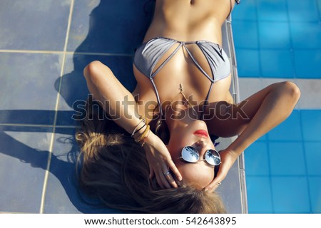 Young beautiful woman in sexy bathing suit with long blonde hair relaxing near pool villa on the tropical beach,red lips,reflect sunglasses,tan skin,sunbathing with coconut oil and cream,enjoy summer