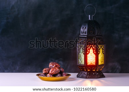 Holy month of Ramadan concept. Burning, lighting, glowing Ramadan Lantern, dates on white table on the background of a dark black gray grey textured wall. Greetings card. Place for text on the left.