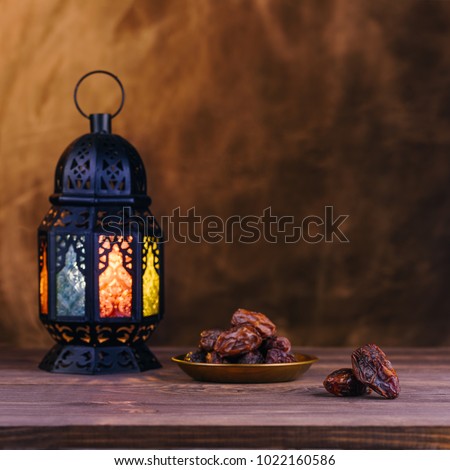 Ramadan concept. Dates close-up in the foreground. On the distant plan a blurry Ramadan Lantern on a wooden table. Textured yellow wall background. Space for text on the right. Square 1:1 frame.