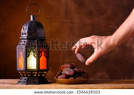 Holy month of Ramadan concept. Righteous Muslim Lifestyle. Fasting. Ramadan lantern, dates. A man\'s hand reaches out to a plate with dates on a wooden table. Dark brown background.
