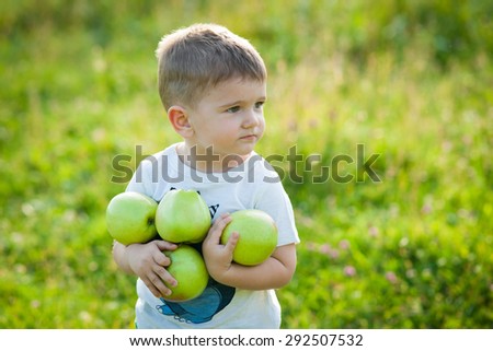 Happy little boy laughs and walks outdoors and holding big green apples. Child biting and eating a large green apple in the garden.