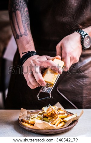 Chief grating parmesan on potato fries in a leather apron, tattooed hands