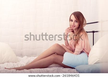 The beautiful young girl in a pink jacket smiles, holding a hand at a mouth, sitting on a bed