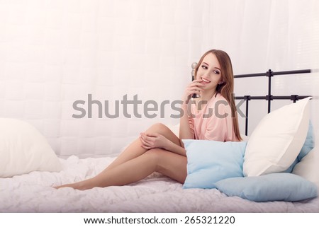 The beautiful young girl in a pink jacket smiles, holding a hand at a mouth, sitting on a bed, looks aside