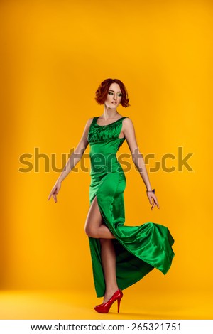 the beautiful red-haired girl in a green dress and red shoes costs, having raised a foot behind on a yellow background, looks aside, having directed hands down