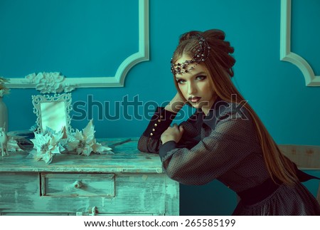 the beautiful young girl with long hair sits at the desktop, one hand on the desktop, another in hair, quietly looks, on a blue background. On the desktop white flowers and the frame for photos.