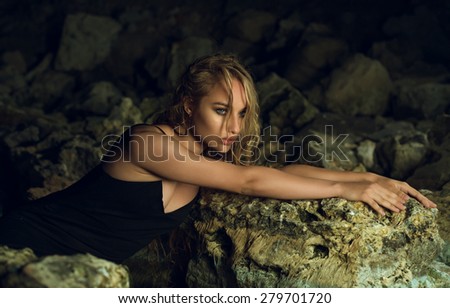 the beautiful young girl with a long fair hair in a black dress sits, having drawn out hands before herself, on stones, looks before herself
