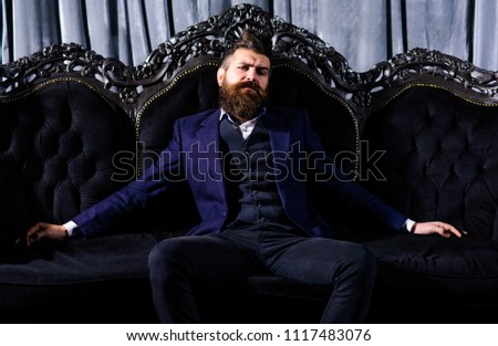 Millionaire in elegant suit sits on luxurious sofa. Bearded man with confident face in classic interior. Luxury lifestyle, confidence, success, rich, power, fashion, wealth, furniture, concept