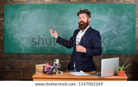 Teacher bearded man tell interesting story. Teacher charismatic hipster stand near table classroom chalkboard background. Teacher interesting interlocutor best friend. Talking to students or pupils.
