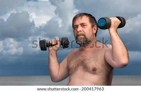 Physical exercises outdoors. Elderly man exercising with dumbbells on sea and cloudy sky background