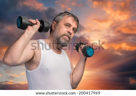 Home Training.  Elderly man exercising with dumbbells on sunset sky background